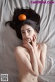 A naked woman laying on a bed with an orange on her head.