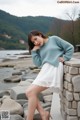 A woman in a blue sweater and white skirt leaning against a stone wall.