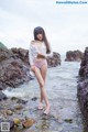 A woman in a white shirt and pink bikini standing on rocks by the water.