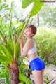 A woman in a white top and blue shorts posing next to a banana tree.